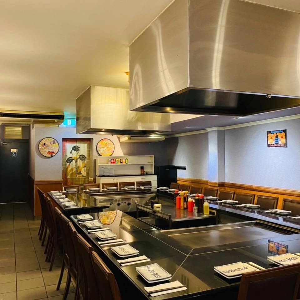 Fujiya’s dining room set for service: two steel teppanyaki hotplates under stainless hoods, ringed by black granite counters and timber chairs