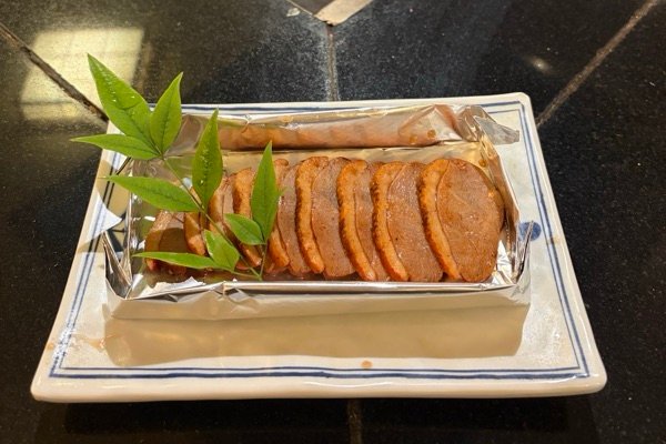Sliced duck breast in special sauce, served in a foil tray on a blue and white plate with a sprig of green leaves