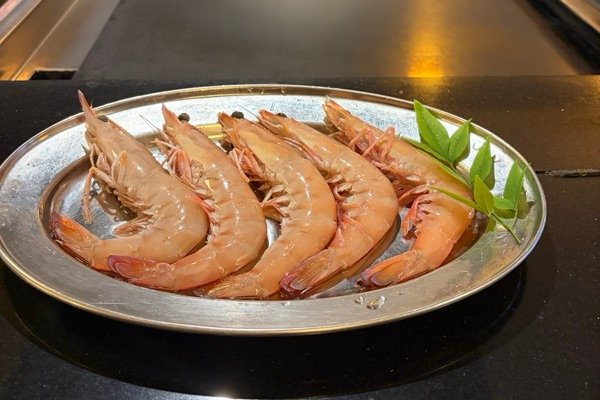 Six raw large king prawns on a silver platter with a sprig of green leaves, in front of the teppanyaki hotplate