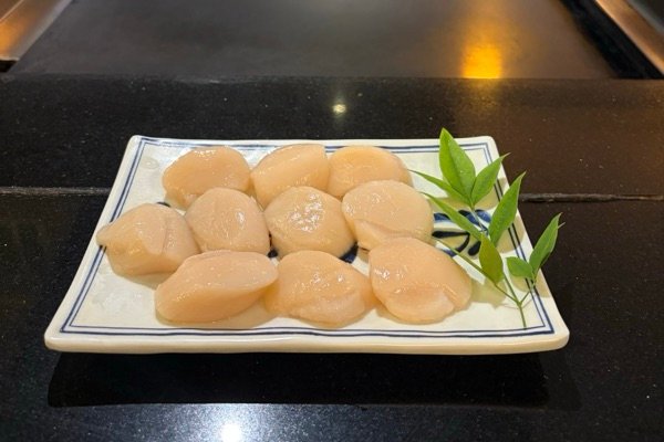 Ten raw Japanese sashimi-grade scallops on a blue and white plate with a sprig of green leaves, in front of the teppanyaki hotplate
