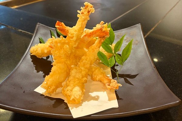 Large king prawn tempura piled on a dark square plate with a sprig of green leaves, at Fujiya’s teppanyaki counter