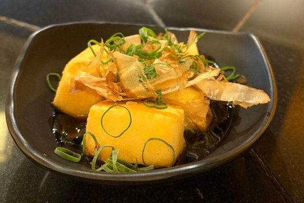 Agedashi tofu in a black bowl, topped with bonito flakes and sliced spring onion