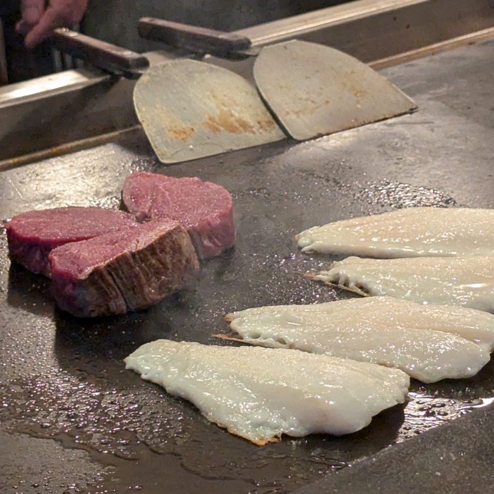 Thick cuts of beef tenderloin and four deep sea perch fillets cooking on the steel teppanyaki hotplate, with the chef’s spatulas resting behind